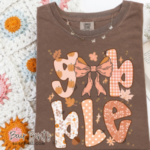 Brown Comfort Colors t-shirt with playful “Gobble” lettering in pumpkin, gingham, floral, and polka dot patterns, featuring a large bow design.