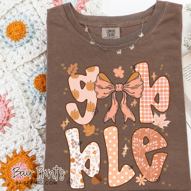 Brown Comfort Colors t-shirt with playful “Gobble” lettering in pumpkin, gingham, floral, and polka dot patterns, featuring a large bow design.