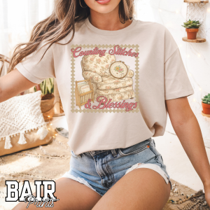 Woman wearing a beige Counting Stitches & Blessings graphic t-shirt featuring a vintage cottagecore chair and cross stitch design, DTG printed cozy craft tee